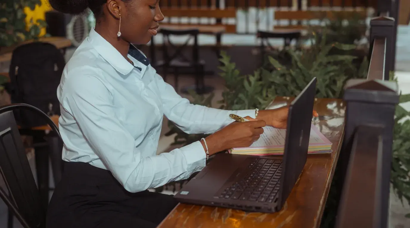 A woman in a pale blue shirt writing on a stack of coloured papers beside an open laptop.