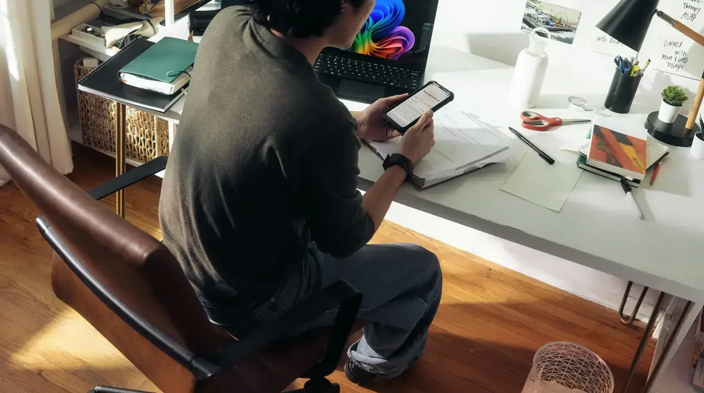Someone seen from behind at a cluttered home desk, checking a phone beside a monitor, loose papers and sticky notes.